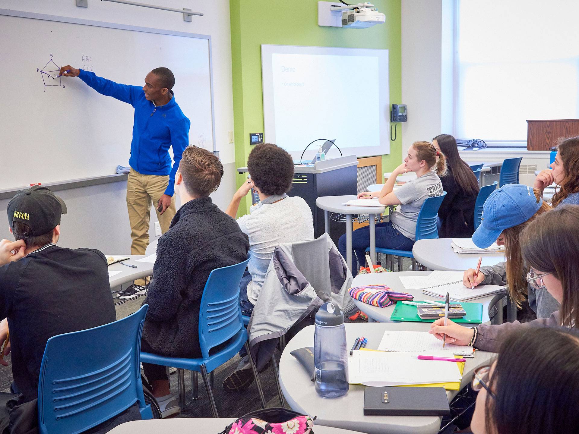 A black student in a blue zip jacket and chinos draws a diagram on a white board in front of his classmates.